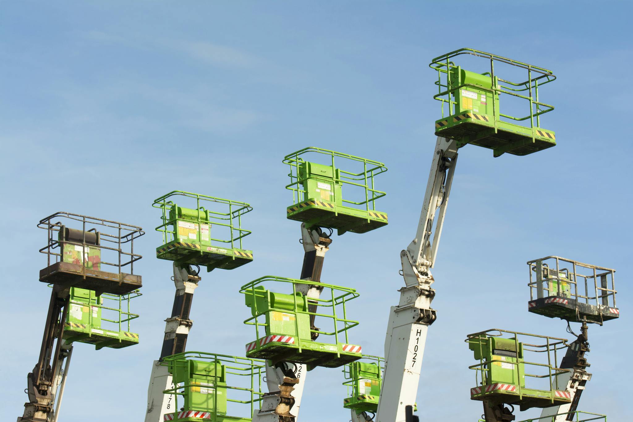 A group of green cherry pickers extending against a clear blue sky in an industrial setting.